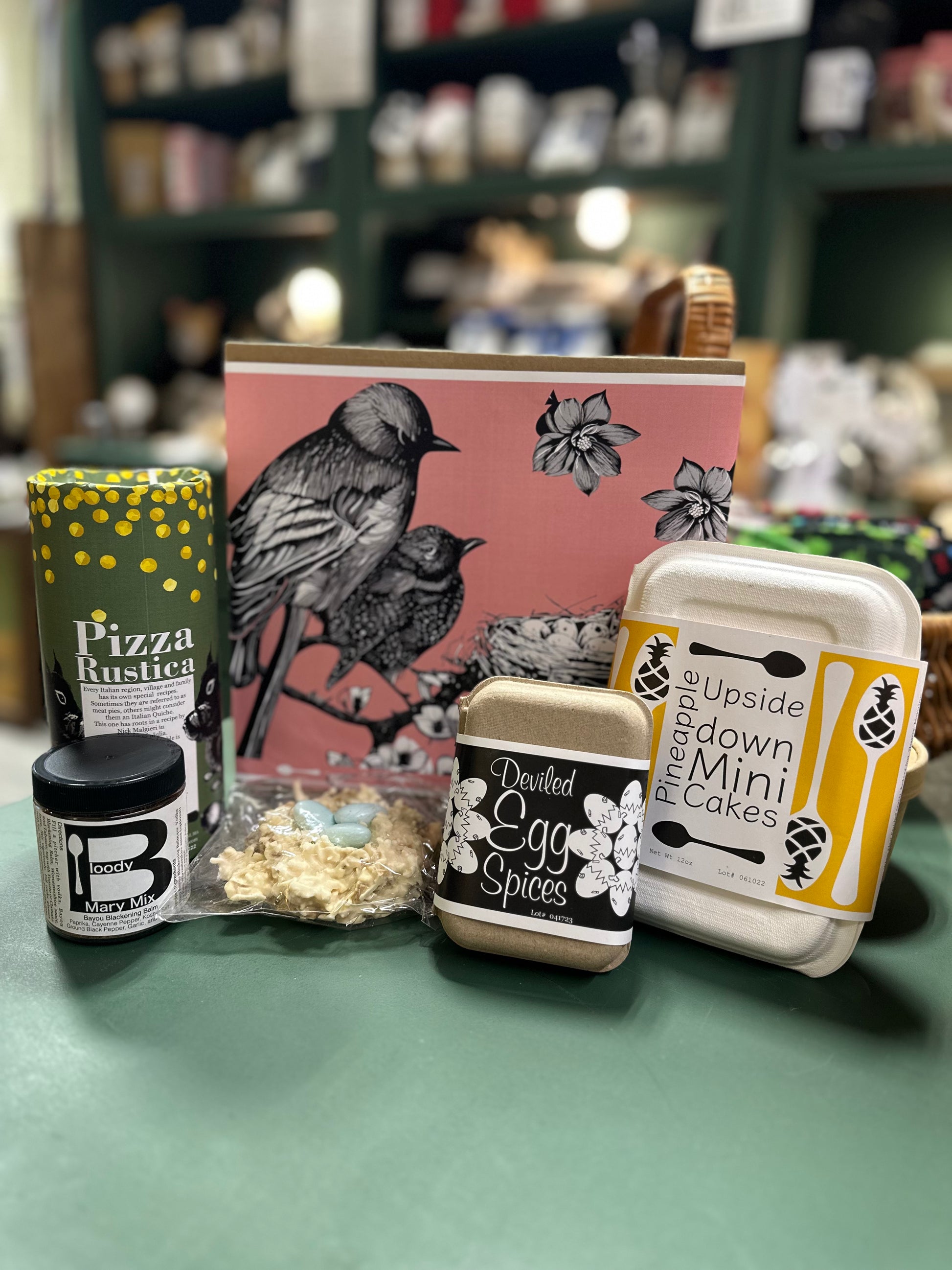A gift box of various food items including Deviled Egg Spices, Bloody Mary Mix, Pizza Rustica, Pineapple Upside-Down Cake, and Birds Nest Bark, displayed on a table with a decorative item in the background.