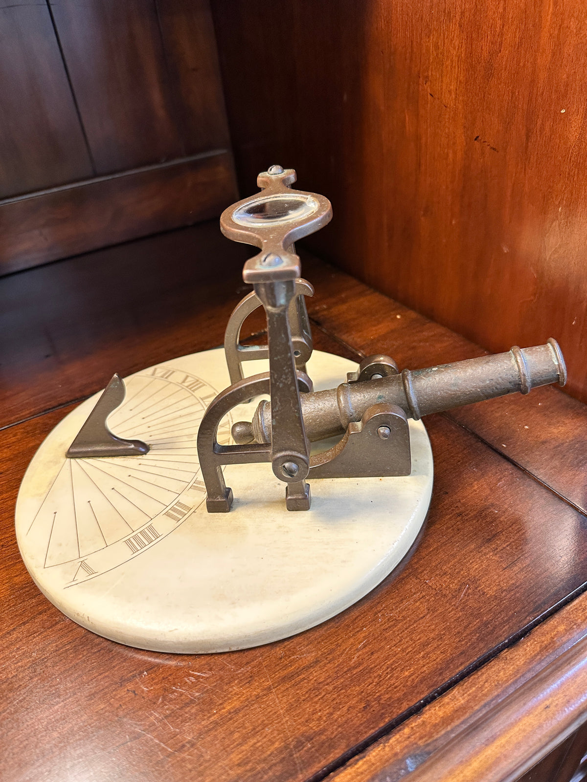 Side view of vintage noon cannon sundial featuring miniature brass cannon, curved metal lens supports and cream-colored base.