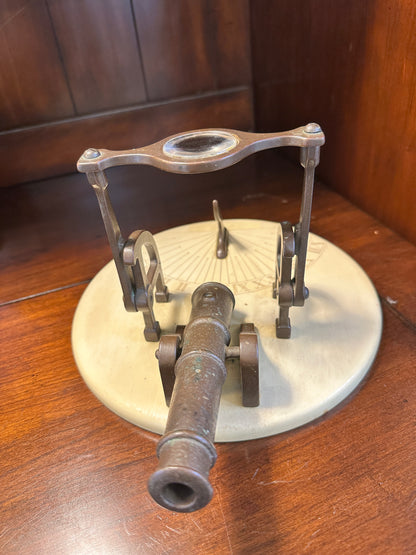 Close-up of vintage miniature brass cannon with open barrel, wheeled carriage and adjustable magnifying glass on circular sundial base.