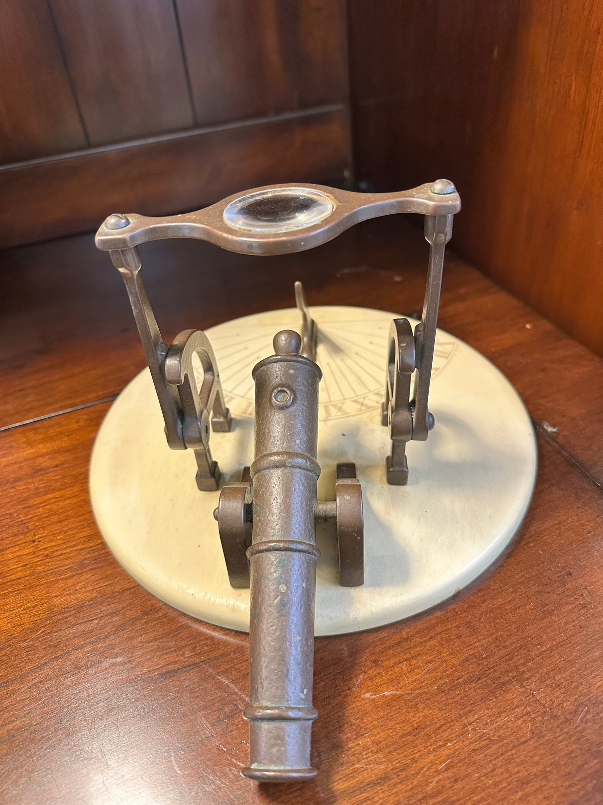 Angled view of vintage brass noon cannon sundial showing miniature cannon barrel beneath raised glass magnifying lens.
