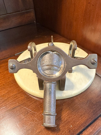 Close-up of vintage brass noon cannon sundial showing chipped magnifying glass, ornate metal supports and cream-colored circular base.